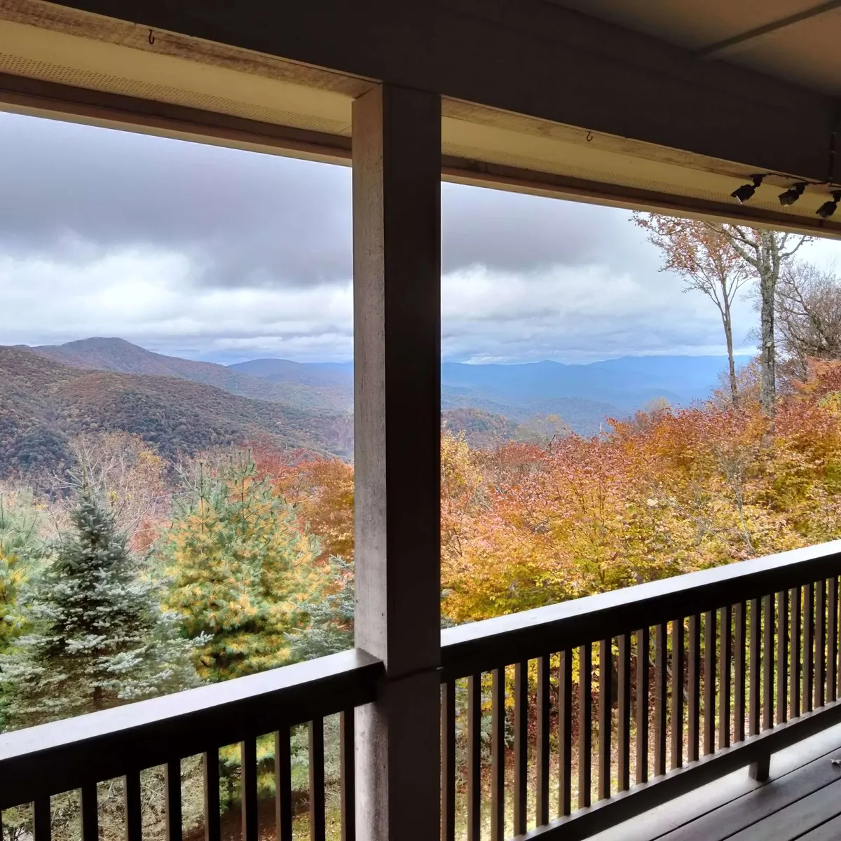 A sweeping view of the Blue Ridge Mountains from a wooden porch, framed by vibrant autumn foliage and a rustic wooden railing. The landscape stretches out into misty peaks, providing a tranquil and immersive mountain experience.