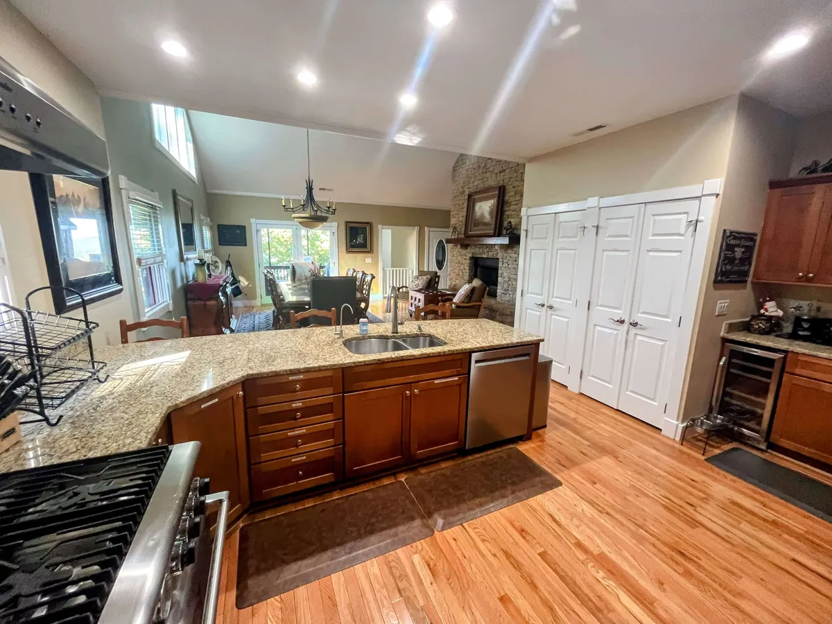 A spacious open-concept interior featuring a modern kitchen with a granite-topped island, high-end stainless steel appliances, and an adjacent dining area. The background reveals a welcoming living space anchored by a fireplace, all bathed in natural light from large windows.