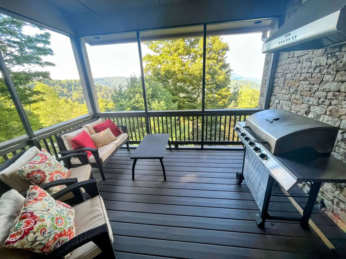 An outdoor dining area is set up on the porch, featuring a comfortable table and chairs under cover. A grill stands nearby, offering the perfect blend of al fresco dining and panoramic views, with the Blue Ridge Mountains in the background.