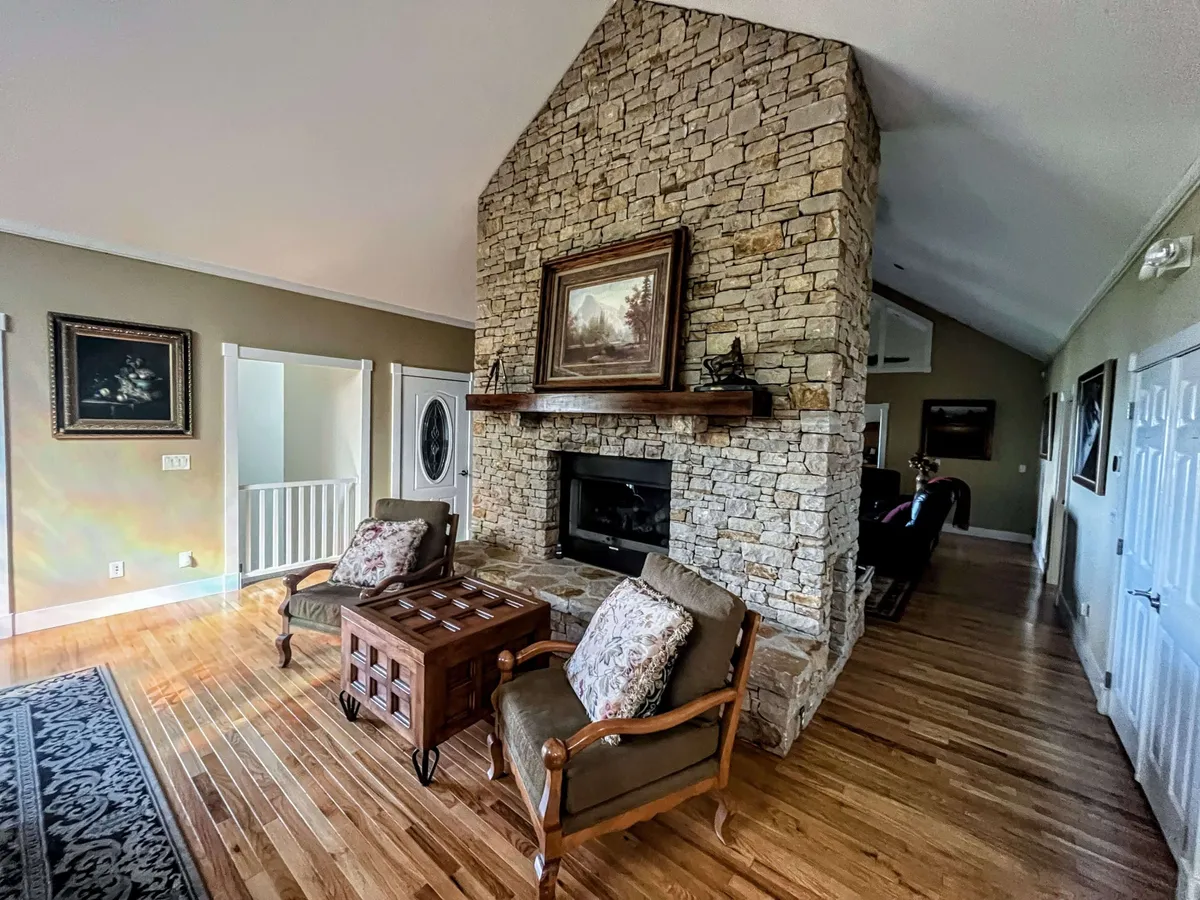 A warm and welcoming living room with a striking stone fireplace, exposed wooden beams, plush sofas, and thoughtfully placed artwork. Large windows allow natural light to pour in, creating a perfect spot for relaxation or socializing while enjoying mountain views.