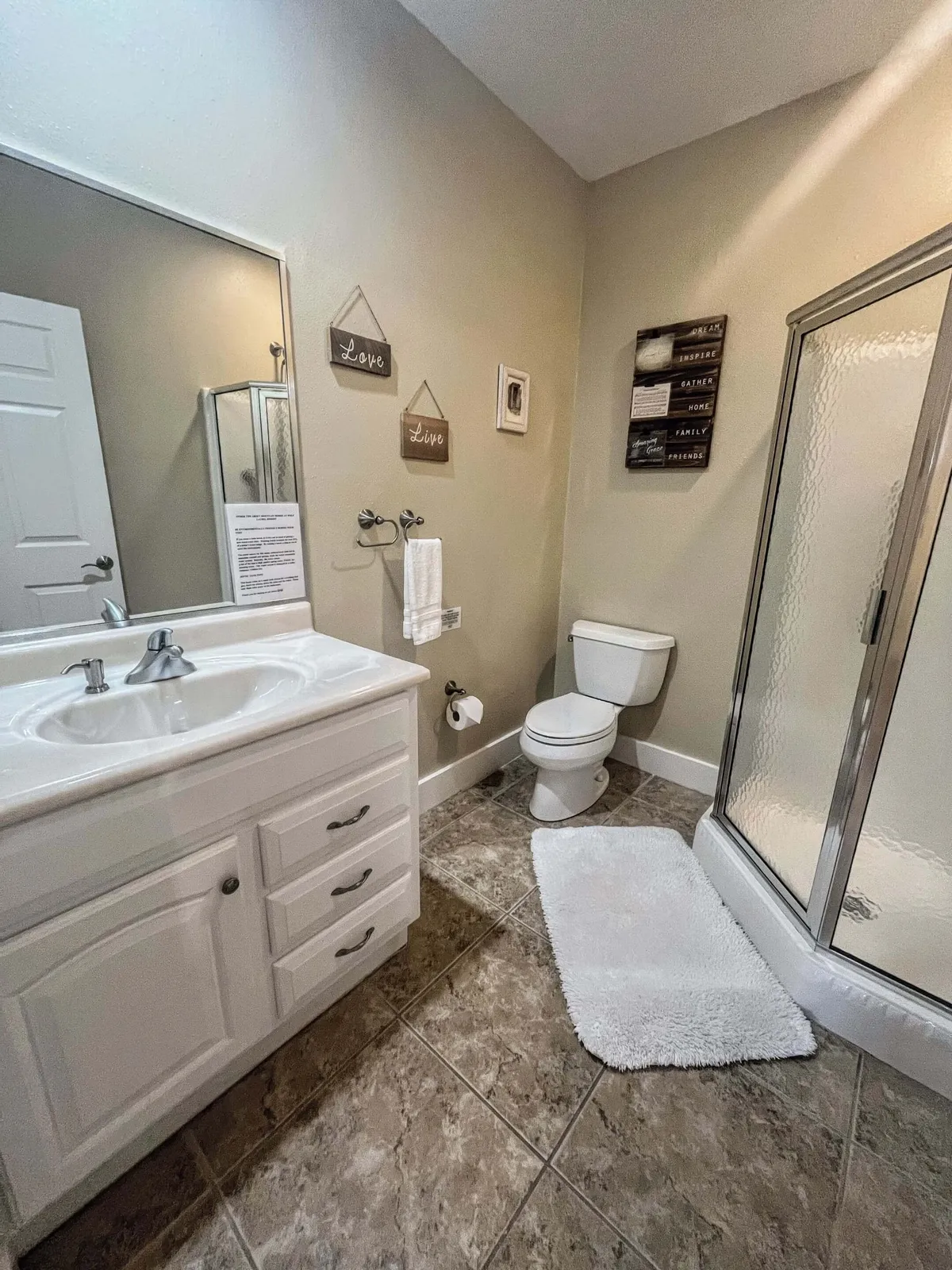 A serene spa-inspired bathroom is shown with a modern vanity and sink, a glass-enclosed shower stall, toilet, and decorative wall art. The lighting is soft, emphasizing the tranquil atmosphere ideal for relaxation and self-care routines.