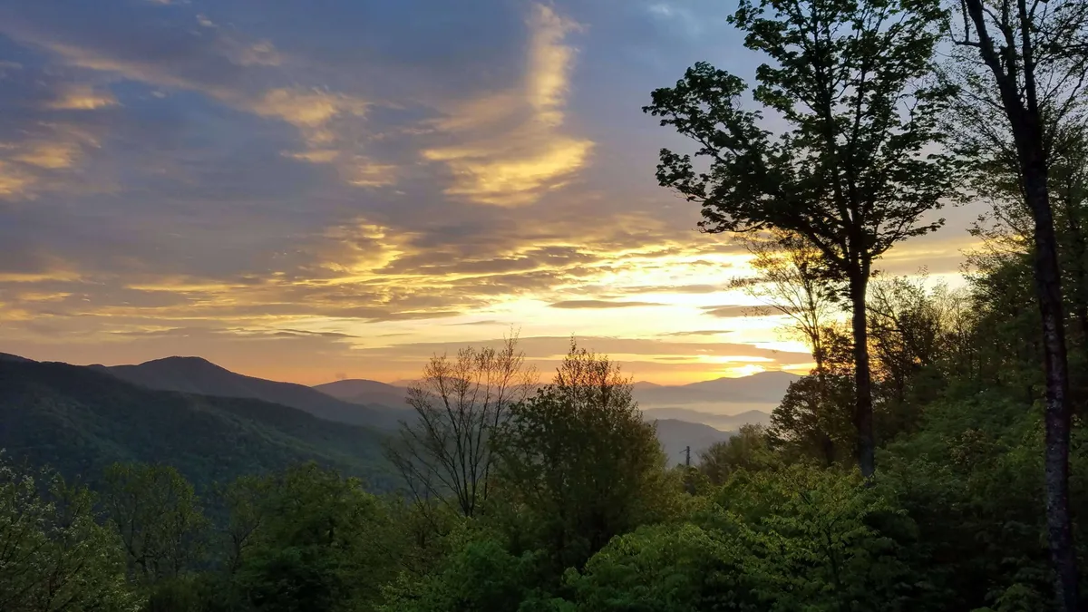 A sweeping mountain range stretches across the horizon with a vivid sunset illuminating the scene, framed by trees in the foreground. The tranquil landscape invites moments of reflection, photography, or simply soaking in the peaceful atmosphere at dusk.