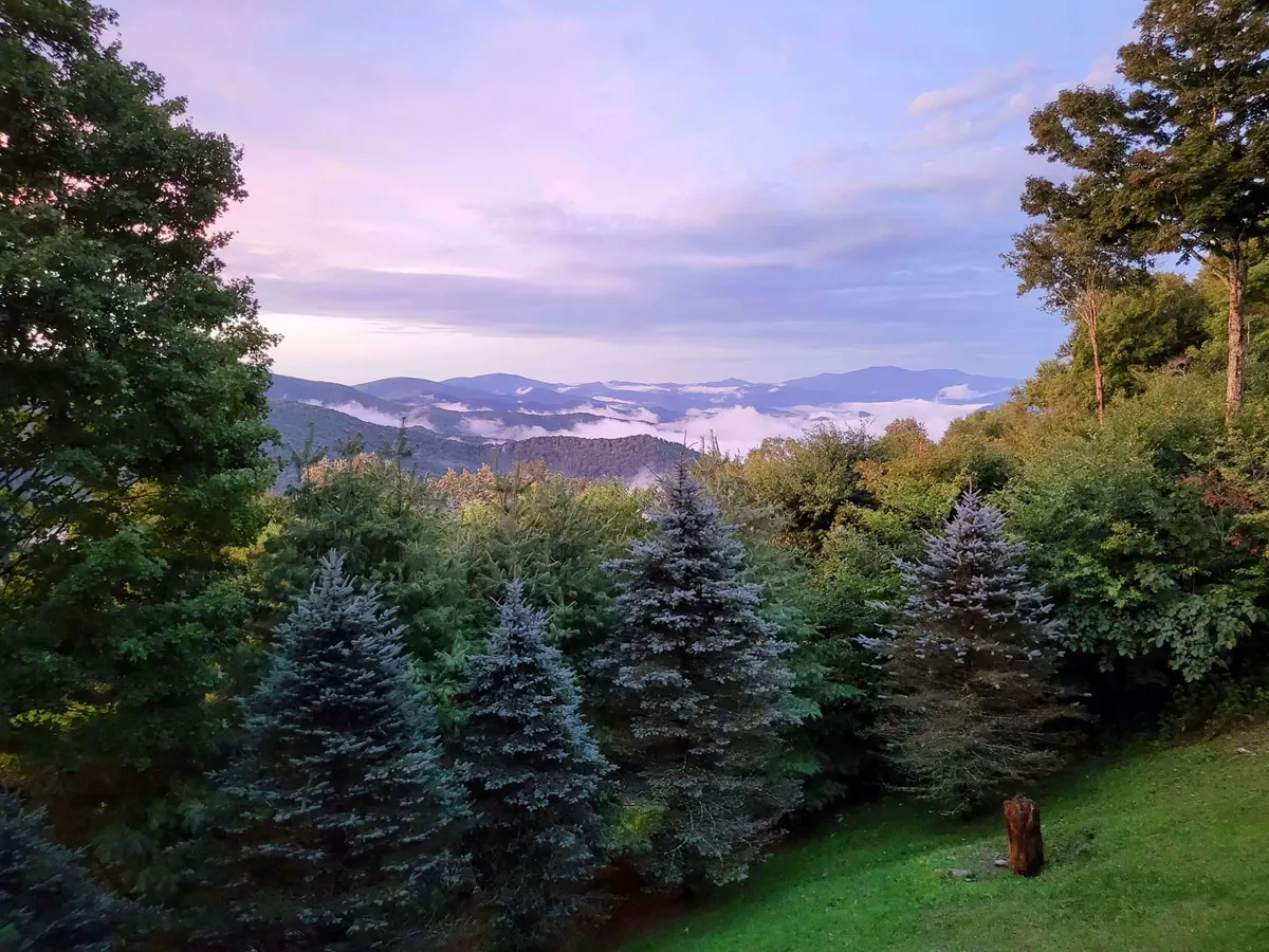 A sweeping view of a mountain range under a sunset sky, framed by green trees and soft clouds. The tranquil atmosphere invites quiet reflection and highlights the beauty of the Blue Ridge landscape.